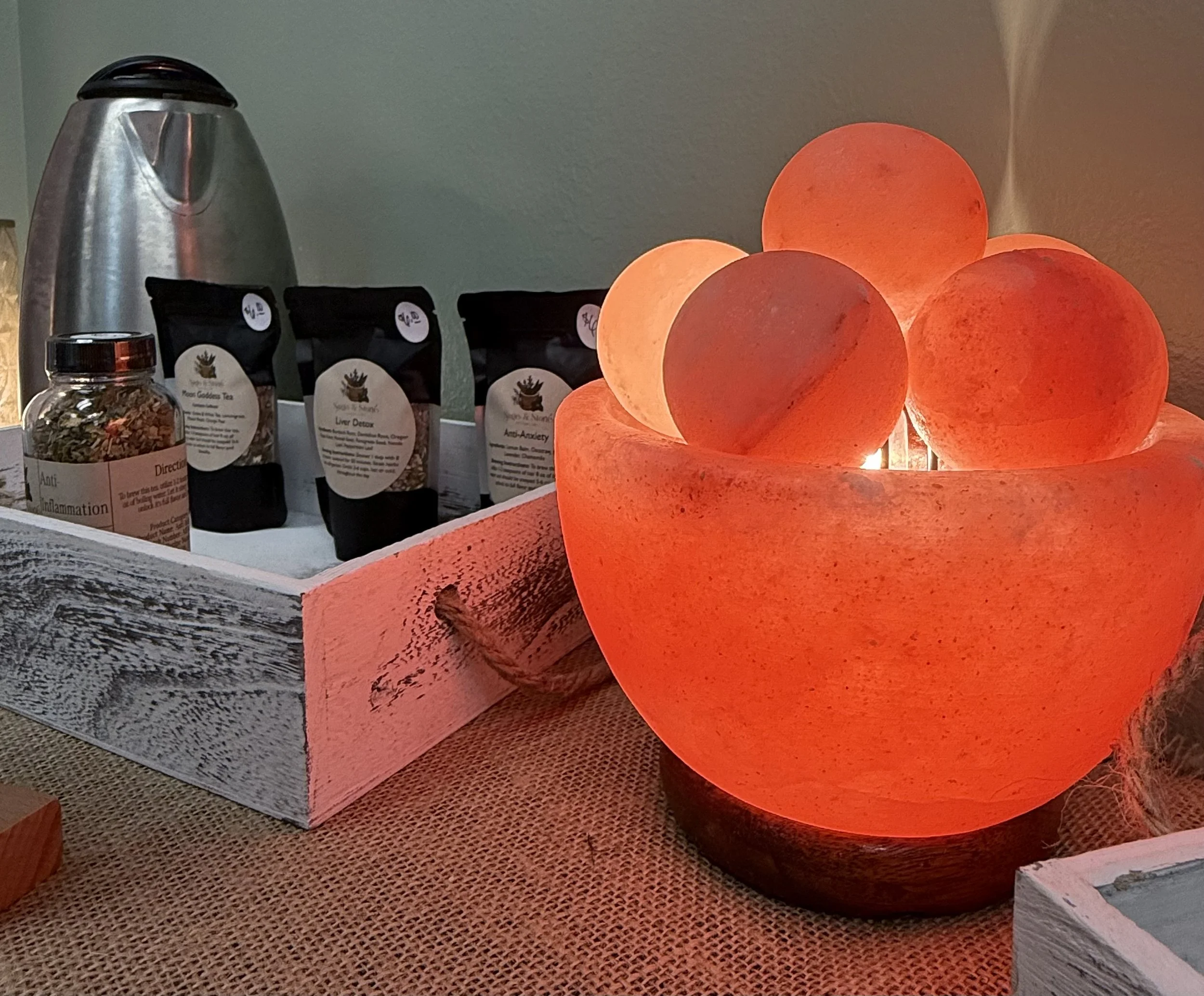 Decorative Himalayan salt lamp shaped like a large sphere with smaller spheres, glowing orange-red. Next to the lamp are bottles of herbal tea and a silver thermos.
