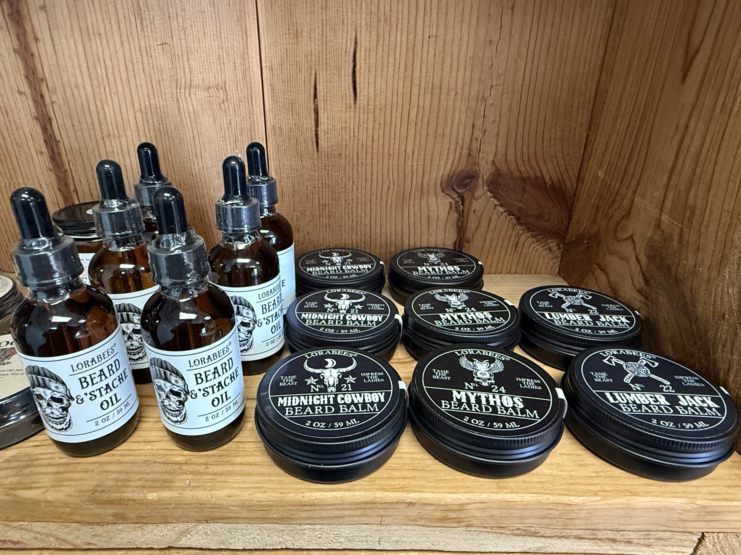 Assorted beard grooming products, including beard oil and beard balm, arranged on a wooden shelf with a wooden wall backdrop.