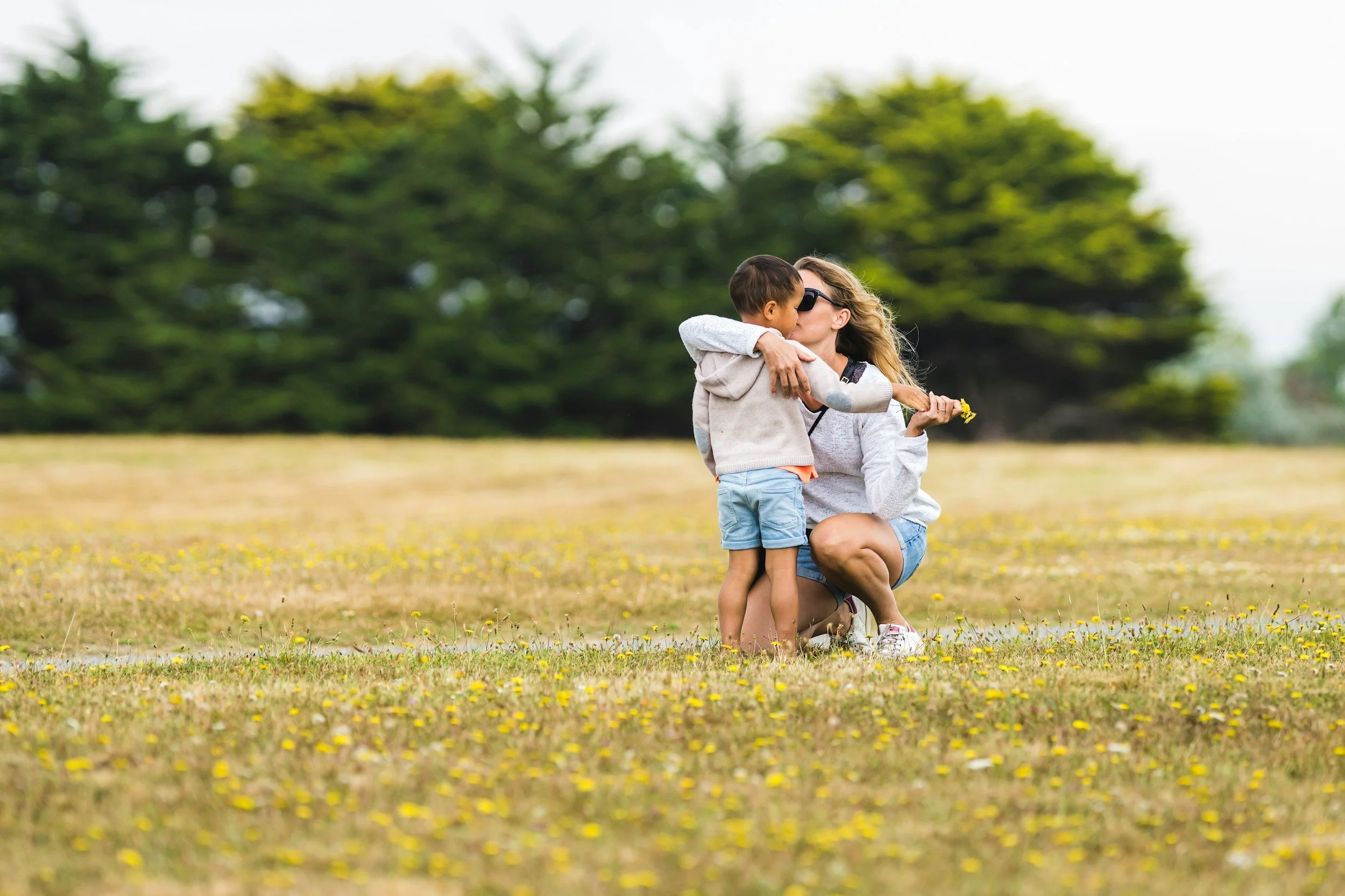 A woman crouching in a field of yellow flowers, holding a young boy and giving him a kiss on the cheek. The boy is holding a yellow flower and wearing sunglasses. The background is filled with lush green trees.