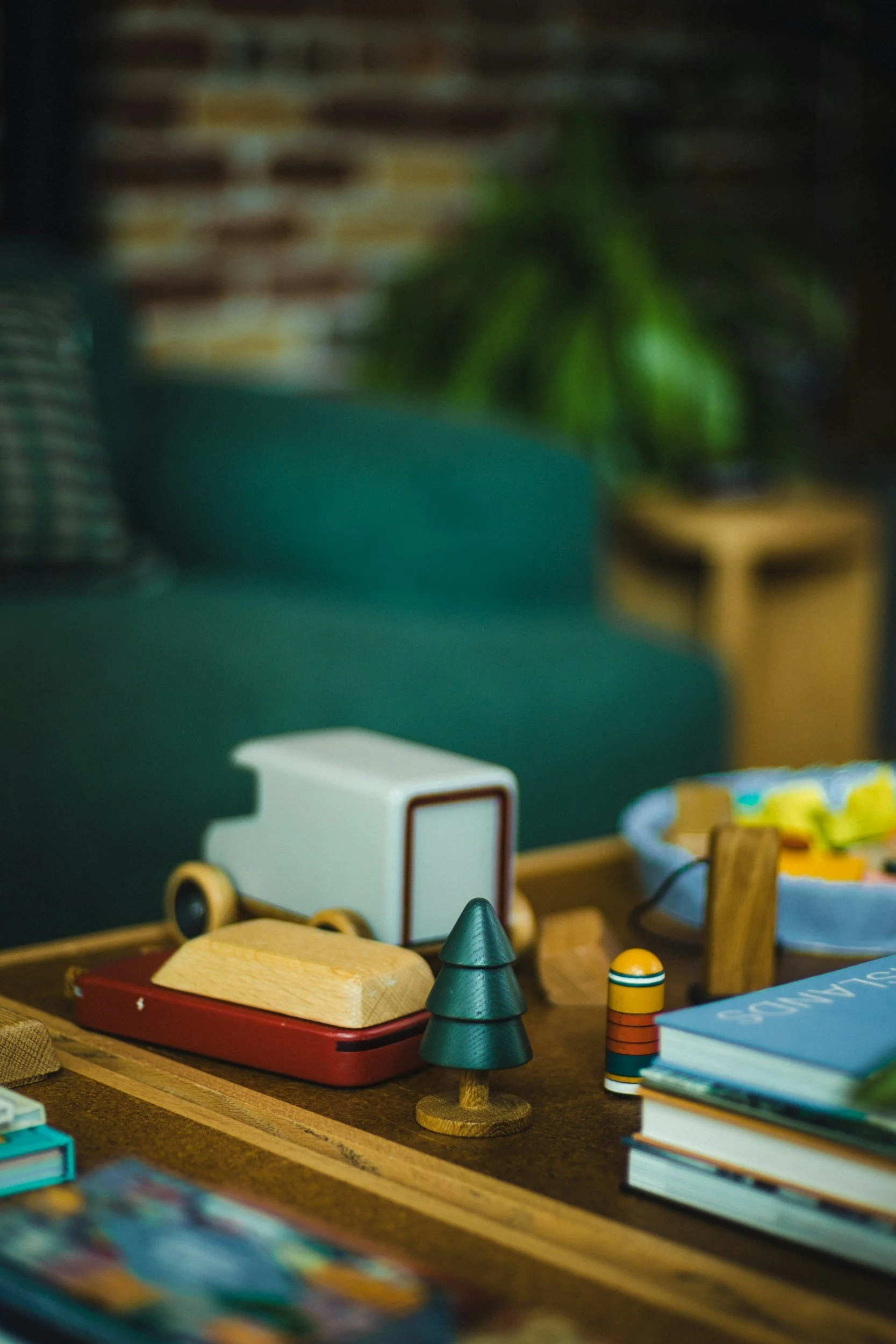 Close-up of a tabletop with various wooden toys, including a small wooden truck, a red boat, green conical trees, and a stack of books, with a blurred background.
