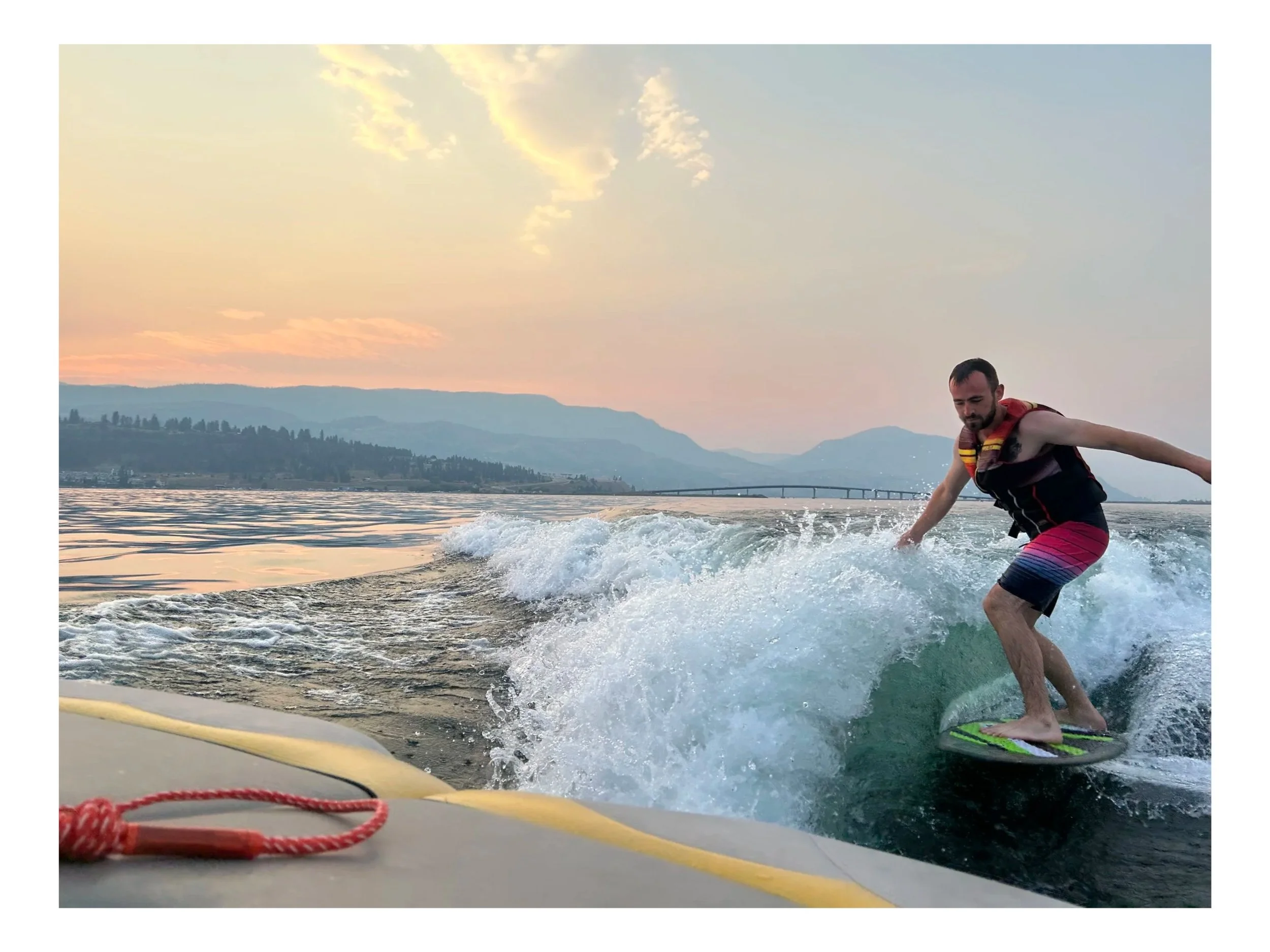 A man water skiing on a lake during sunset, wearing a life jacket and colorful shorts, with mountains and a bridge in the background.
