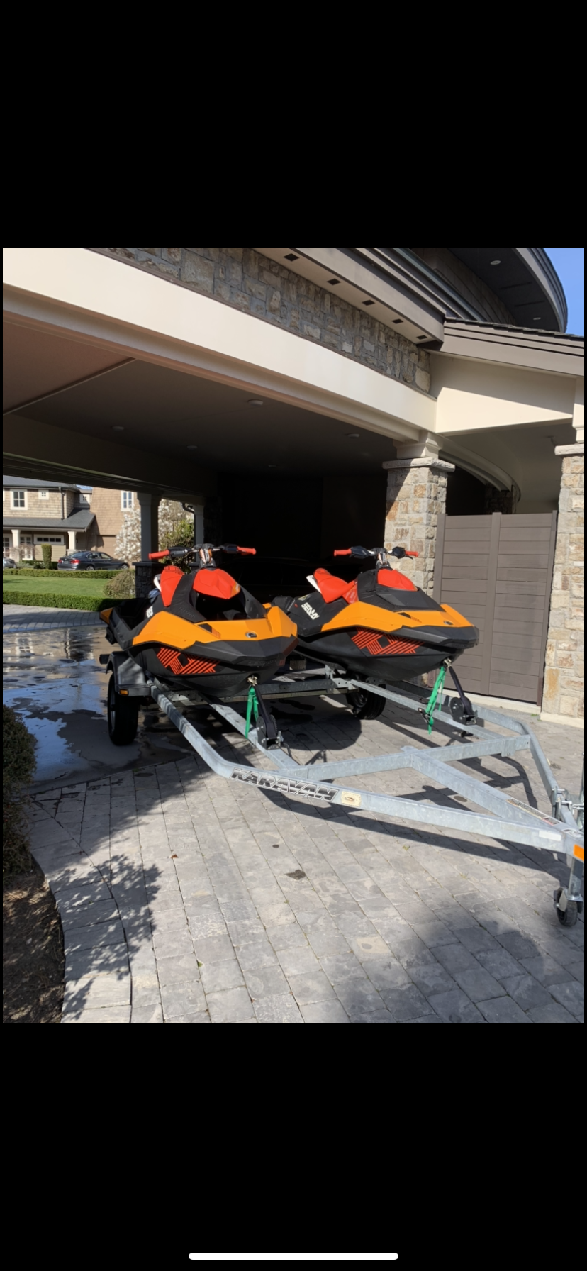 Two orange and black snowmobiles on a trailer parked outside a house with a stone and stucco exterior.