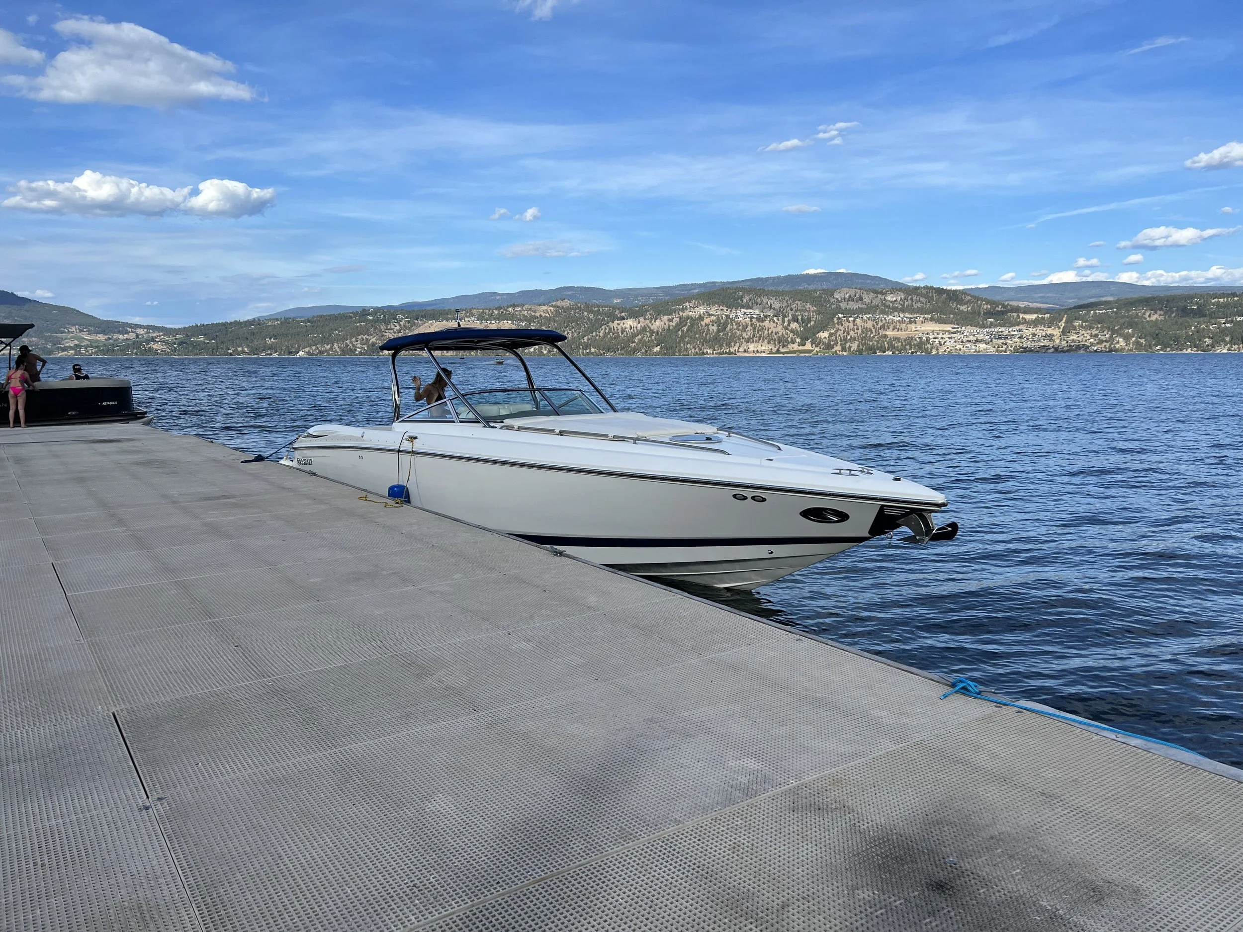 A dock by a lake with a large white motorboat docked and small people nearby, with hills and a blue sky with clouds in the background.
