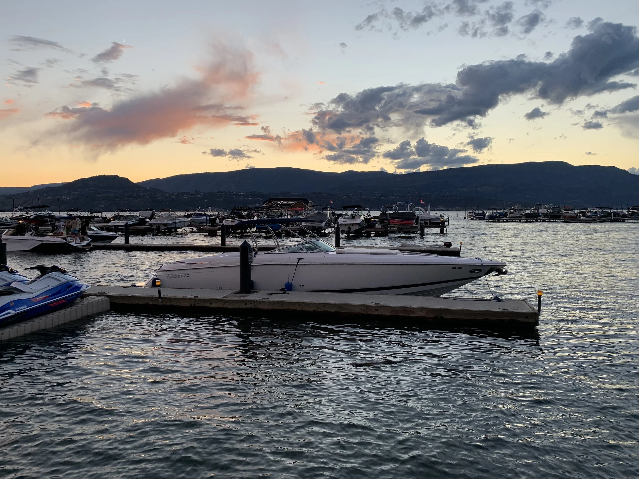Marina with boats docked on a lake at sunset with a mountain range in the background and cloudy sky.