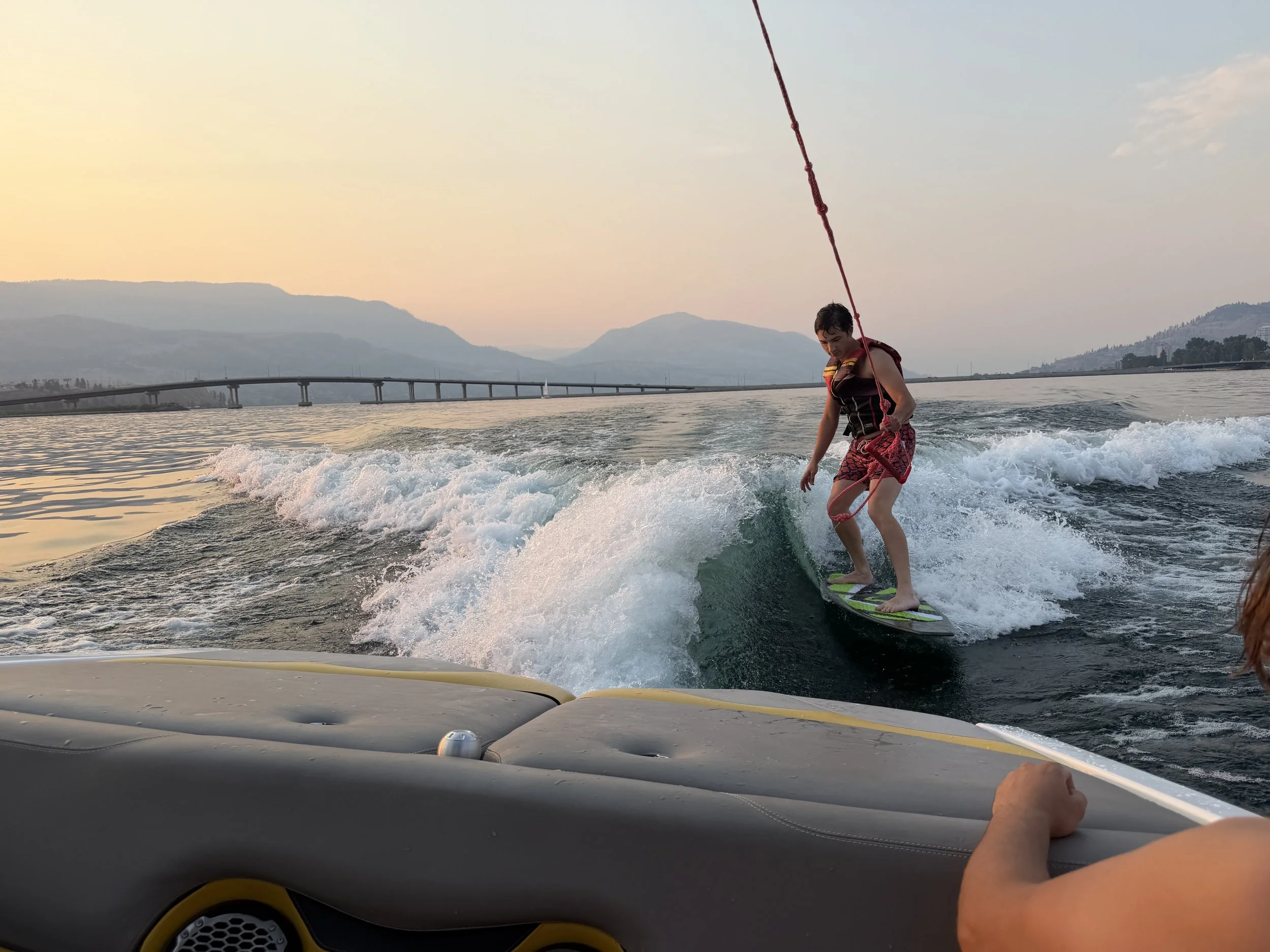 A person wakeboarding on a lake at sunset, with mountains and a bridge in the background, seen from inside a boat.