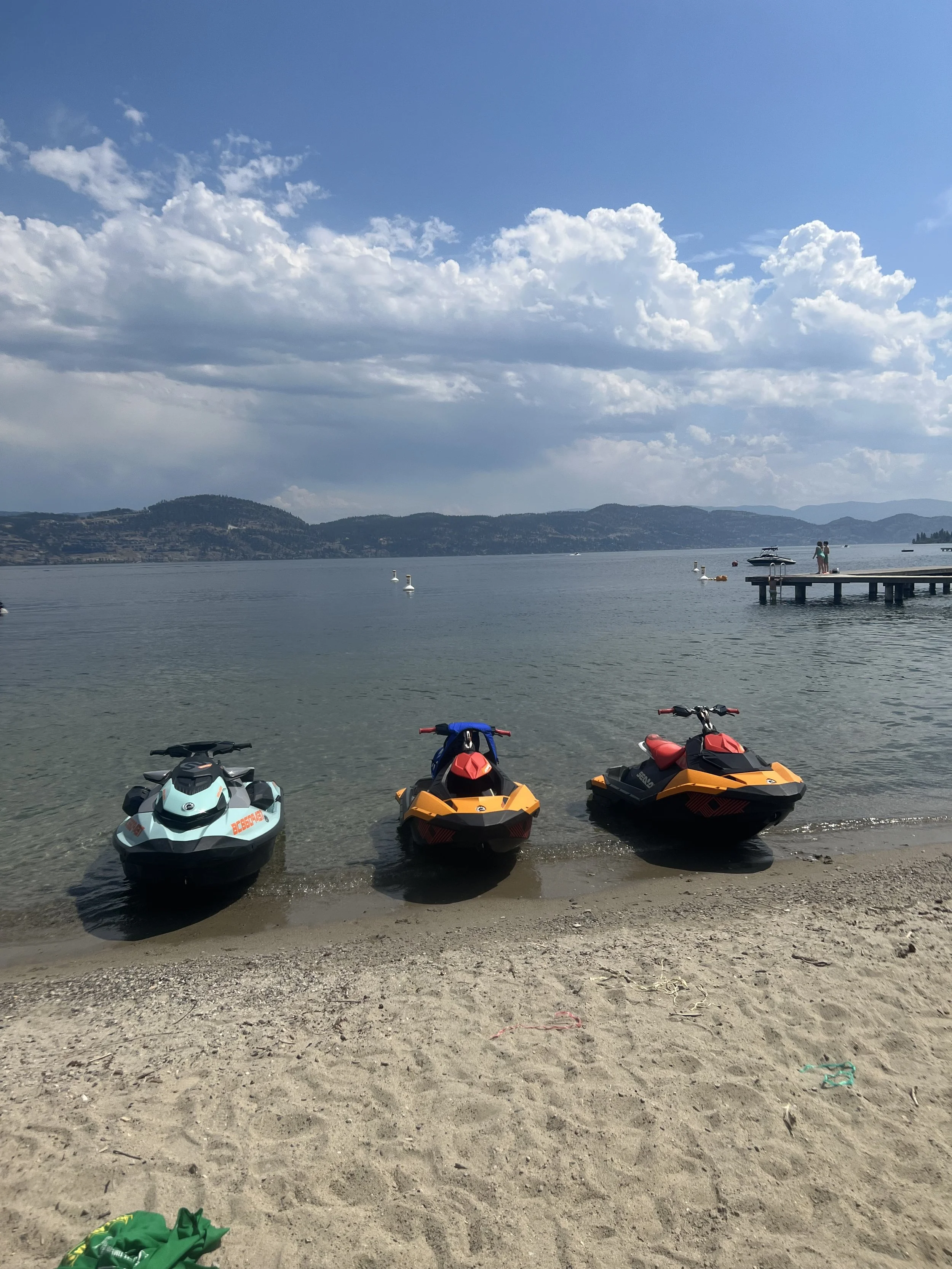 Three personal watercraft (jet skis) parked on the sandy shore of a large lake, with a pier and boats in the distance, under a partly cloudy sky with mountains in the background.