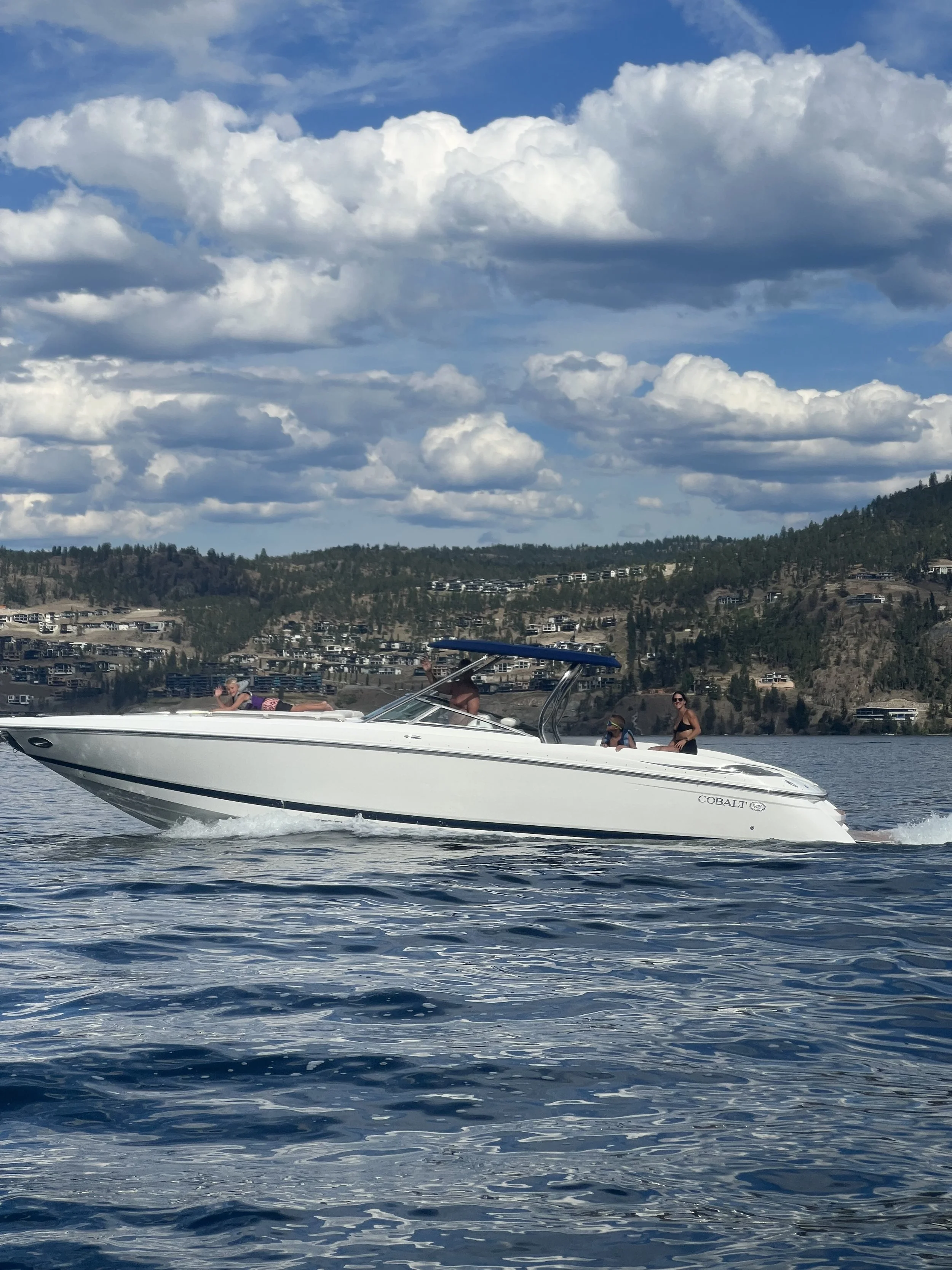 A white motorboat on a lake with a person lying on the bow, two people sitting inside, and two women standing at the stern. The backdrop features a hillside with residential buildings and a sky filled with large white clouds.