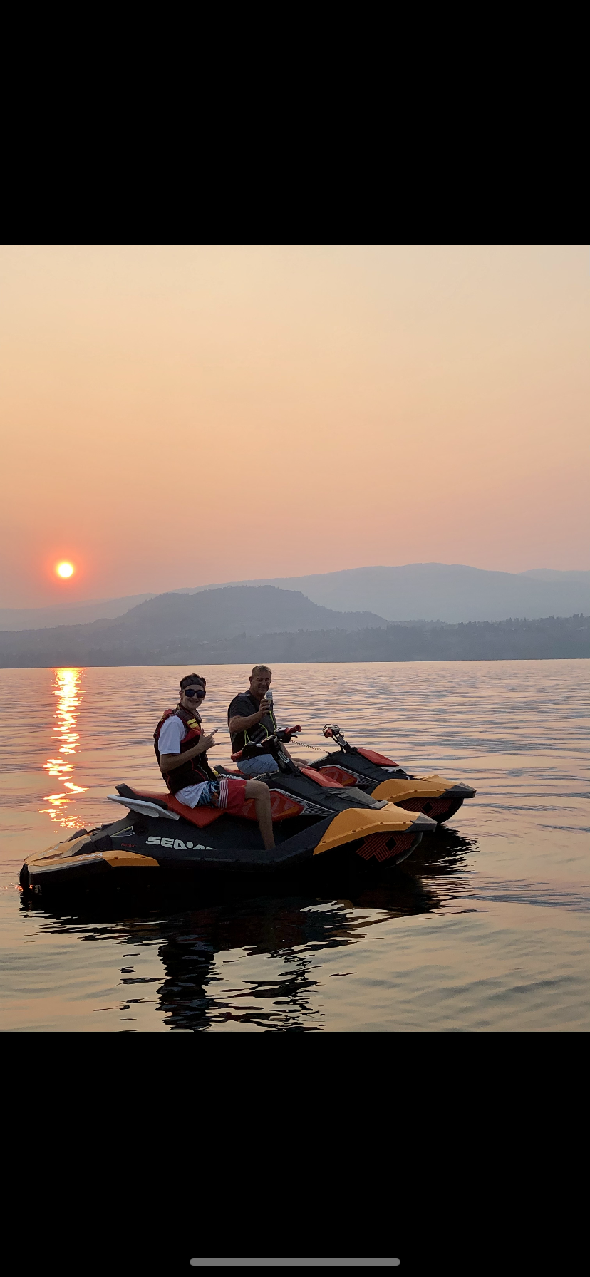 Two men riding jet skis on a calm lake during sunset, with mountainous landscape in the background.