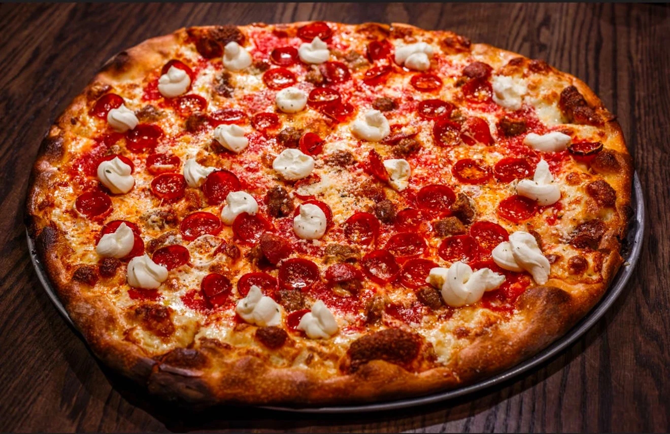 A whole pepperoni pizza with dollops of white cheese or sour cream on top, placed on a dark wooden surface.