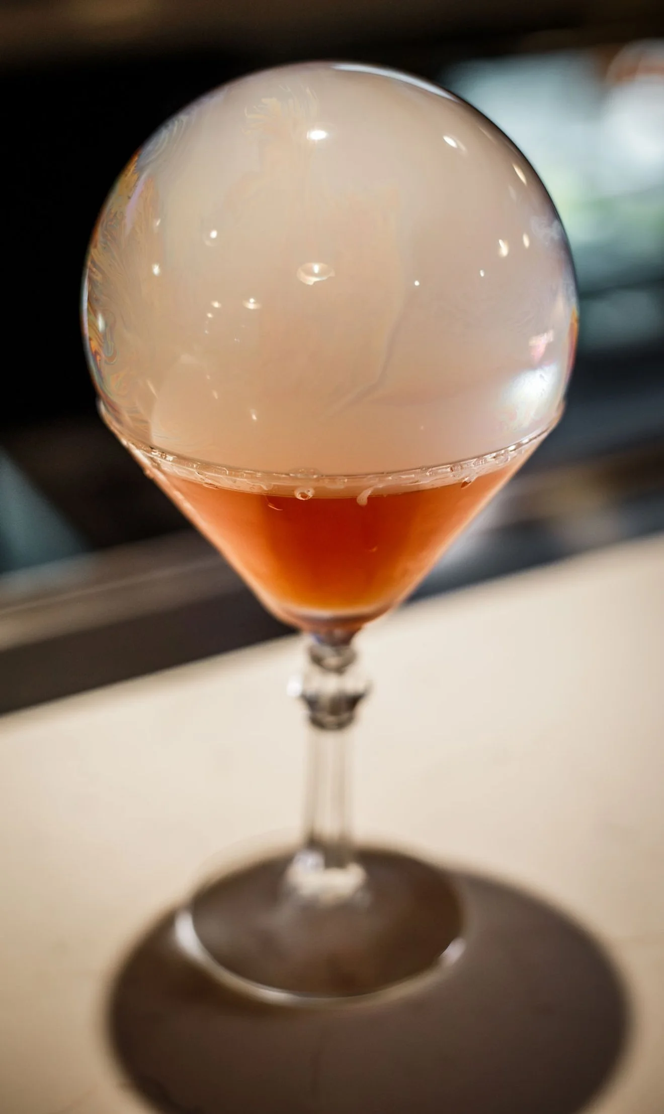 Close-up of a cocktail glass with a layered pink and white drink topped with a large, clear, spherical smoke bubble.