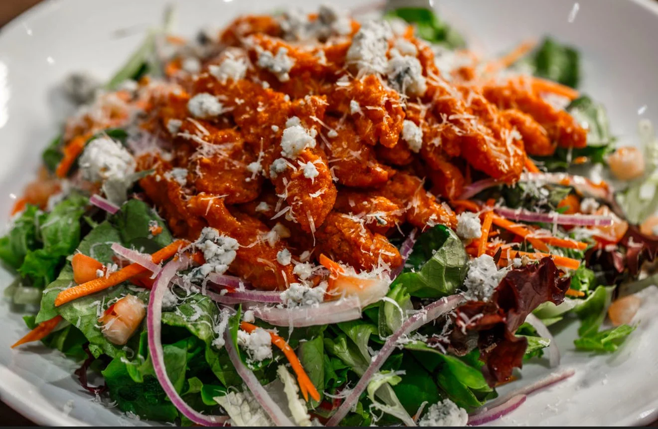 Salad with mixed greens, shredded carrots, red onions, shredded cheese, and pieces of seasoned chicken in a red sauce on top