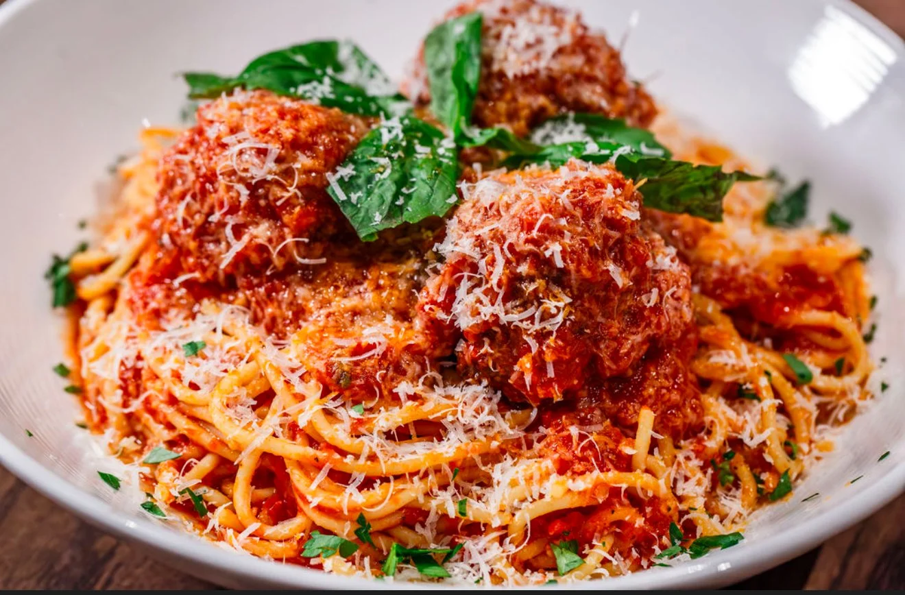 Spaghetti pasta with marinara sauce, meatballs, grated cheese, and fresh basil in a white bowl.