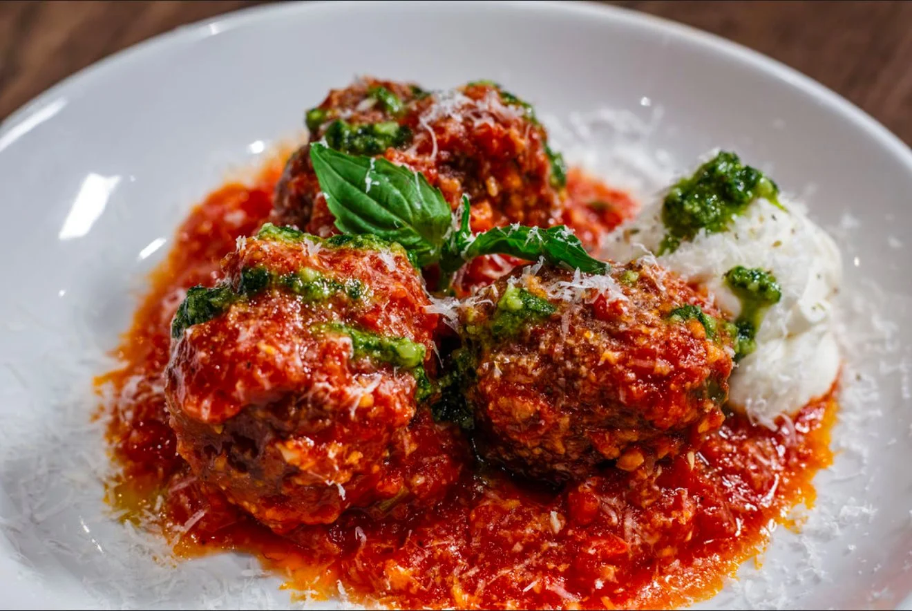 Plate of Italian meatballs with marinara sauce, garnished with basil, served with a side of ricotta cheese and green pesto.