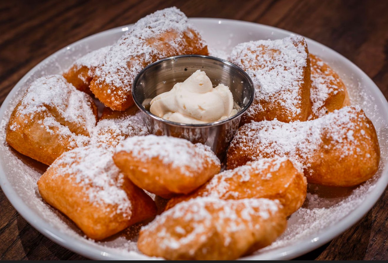 Fried dough squares dusted with powdered sugar on a white plate with a small cup of cream in the center.
