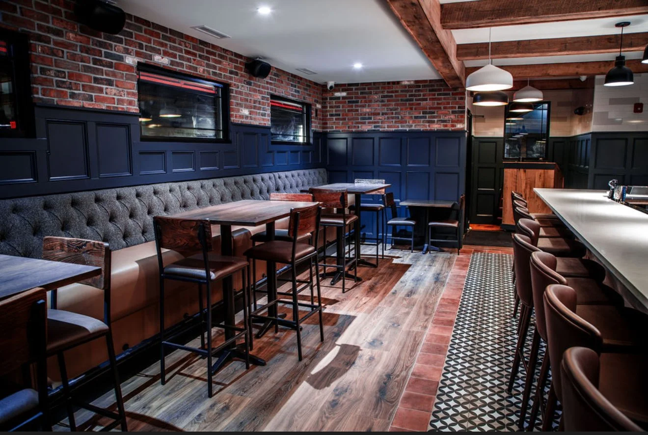 Interior of a modern restaurant with exposed brick walls, dark blue paneled wainscoting, wooden ceiling beams, and a mix of high-top and bar seating with upholstered chairs.