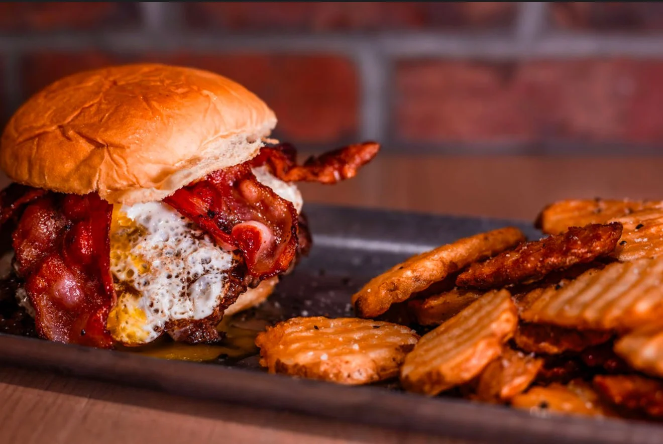Close-up of a bacon cheeseburger with a fried egg, served with seasoned potato chips on a black tray.