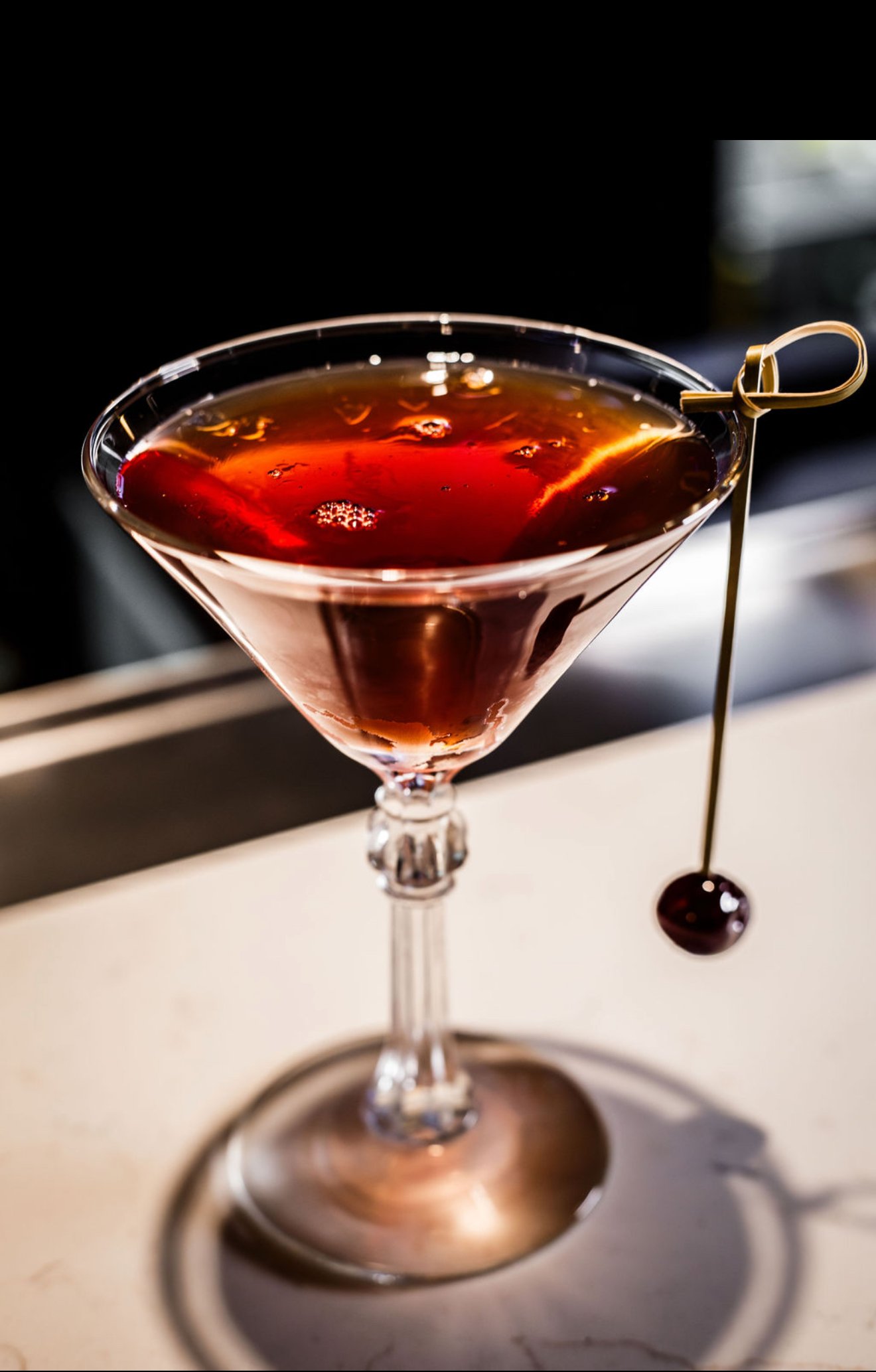 A cocktail glass with a dark red drink, garnished with a cherry on a skewer. The glass has a decorative stem and is set on a bar or counter with a dark background.