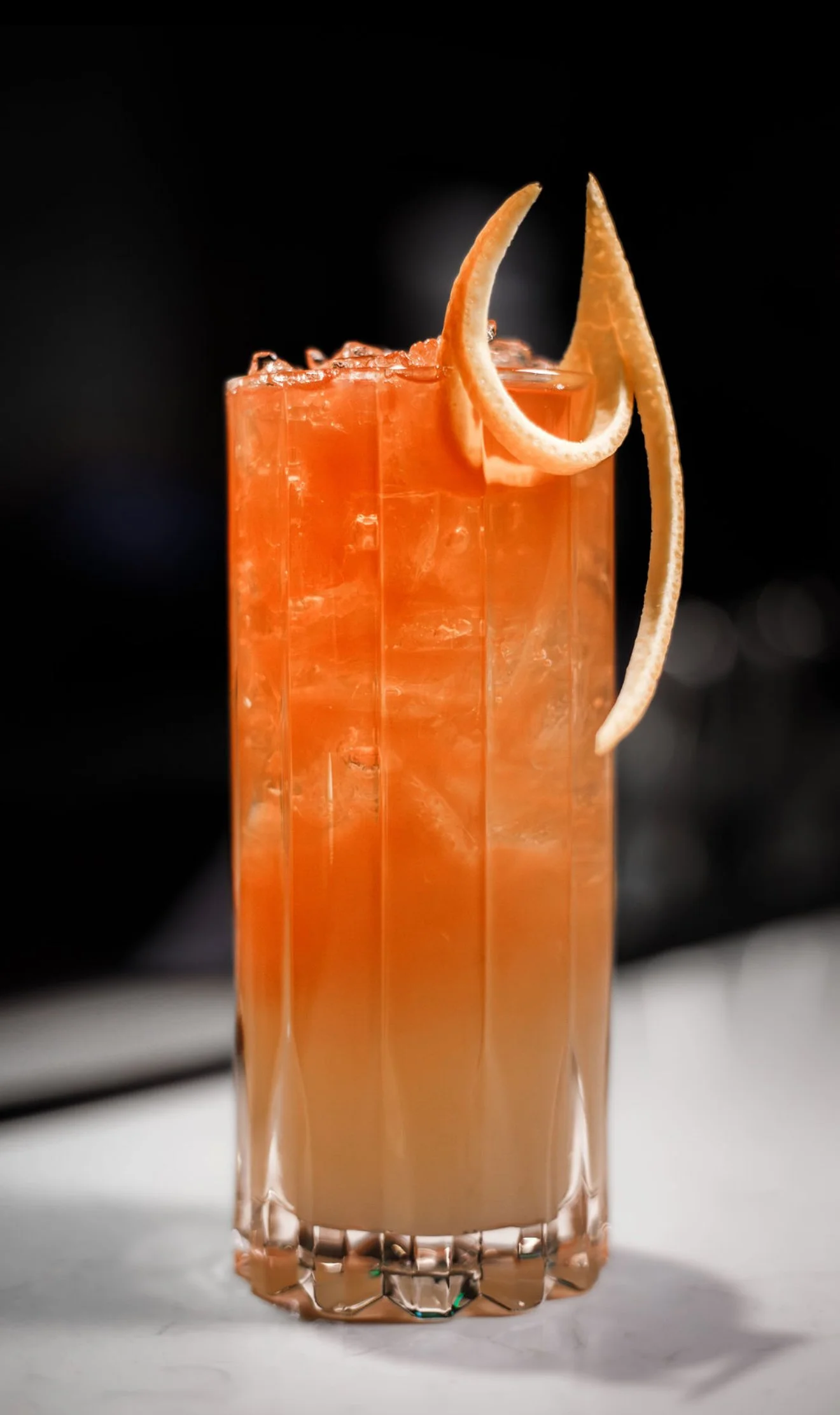 A tall glass of orange cocktail with lemon twists garnishing the top, against a black background.