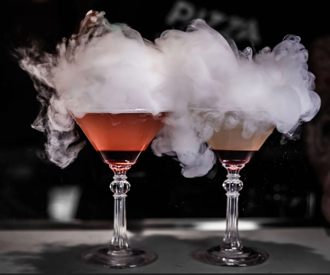 Two martini glasses filled with a pink cocktail, with thick white smoke or vapor emanating from the top of each glass, creating a foggy effect against a dark background.