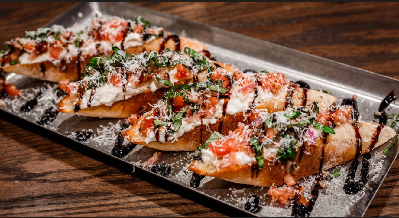 Three slices of pizza on a rectangular black tray, topped with cheese, chopped tomatoes, chopped basil, and drizzled with balsamic glaze, with grated cheese sprinkled on top, placed on a wooden table.
