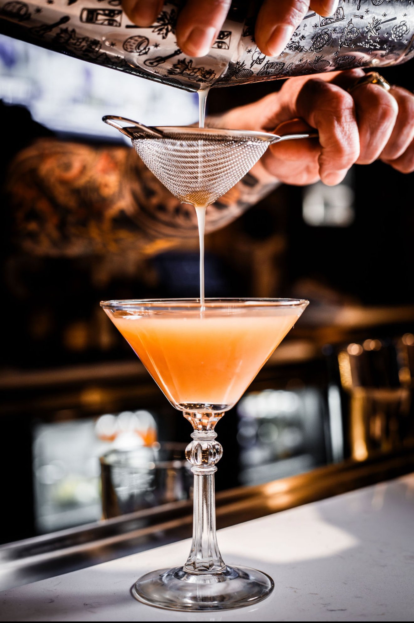 A bartender pouring a pink drink through a fine mesh strainer into a martini glass. The background is blurred, emphasizing the drink and the bartender's hands.