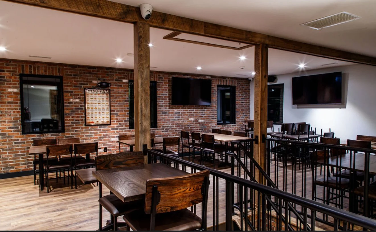 Interior of a restaurant or cafe with wooden tables and chairs, brick walls with windows and TV screens, and a staircase with a black railing.