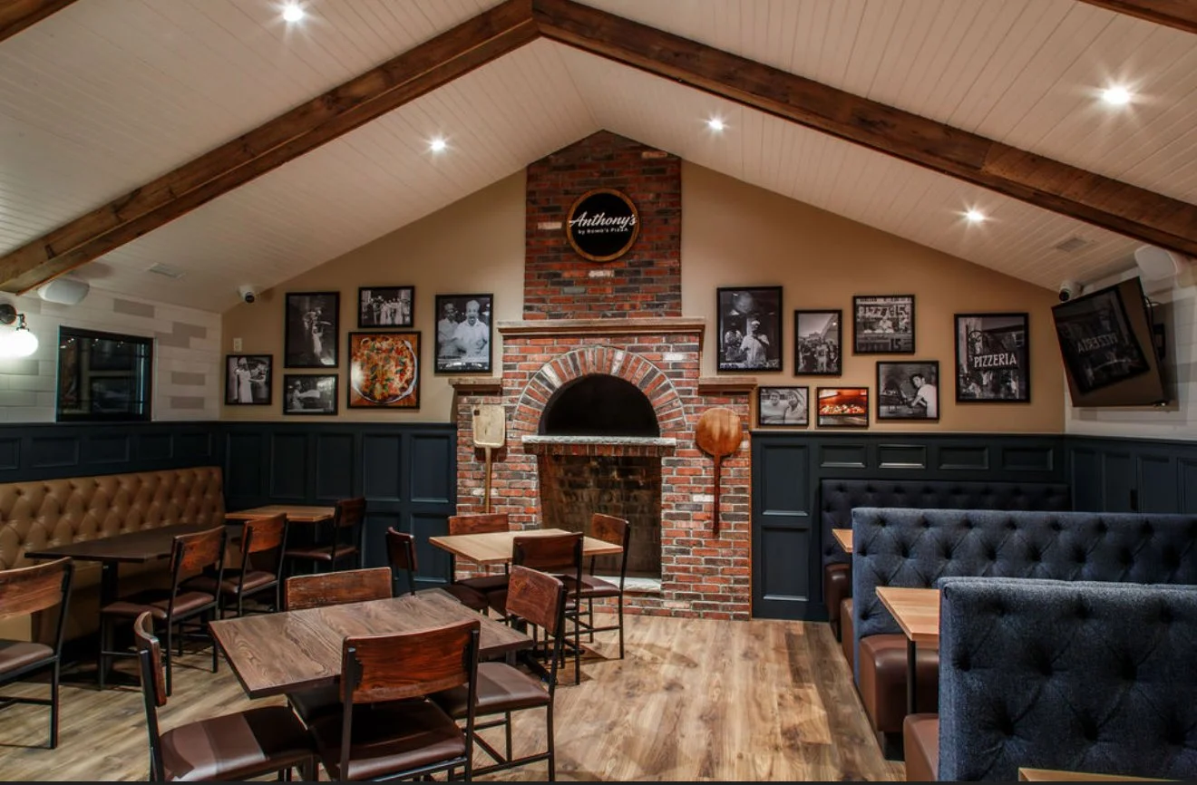 Interior view of a restaurant with wooden tables and chairs, a brick fireplace, framed black-and-white photos on the wall, and a TV mounted on the wall.