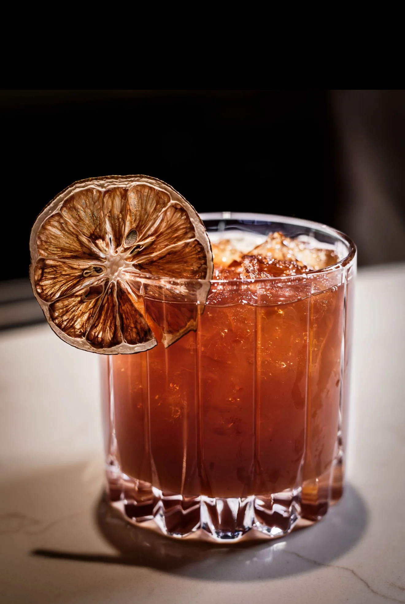 A close-up of a glass filled with a red drink and ice, garnished with a slice of dried lemon on the rim.