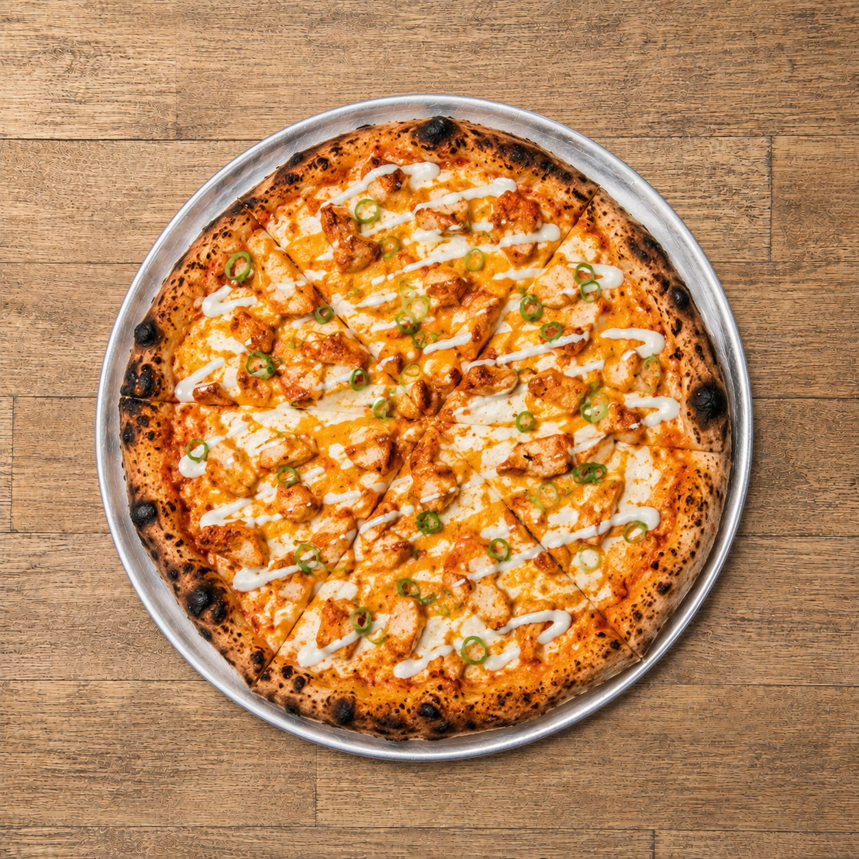 A pizza with chicken, cheese, green onions, and white sauce on a metal tray, placed on a wooden table.