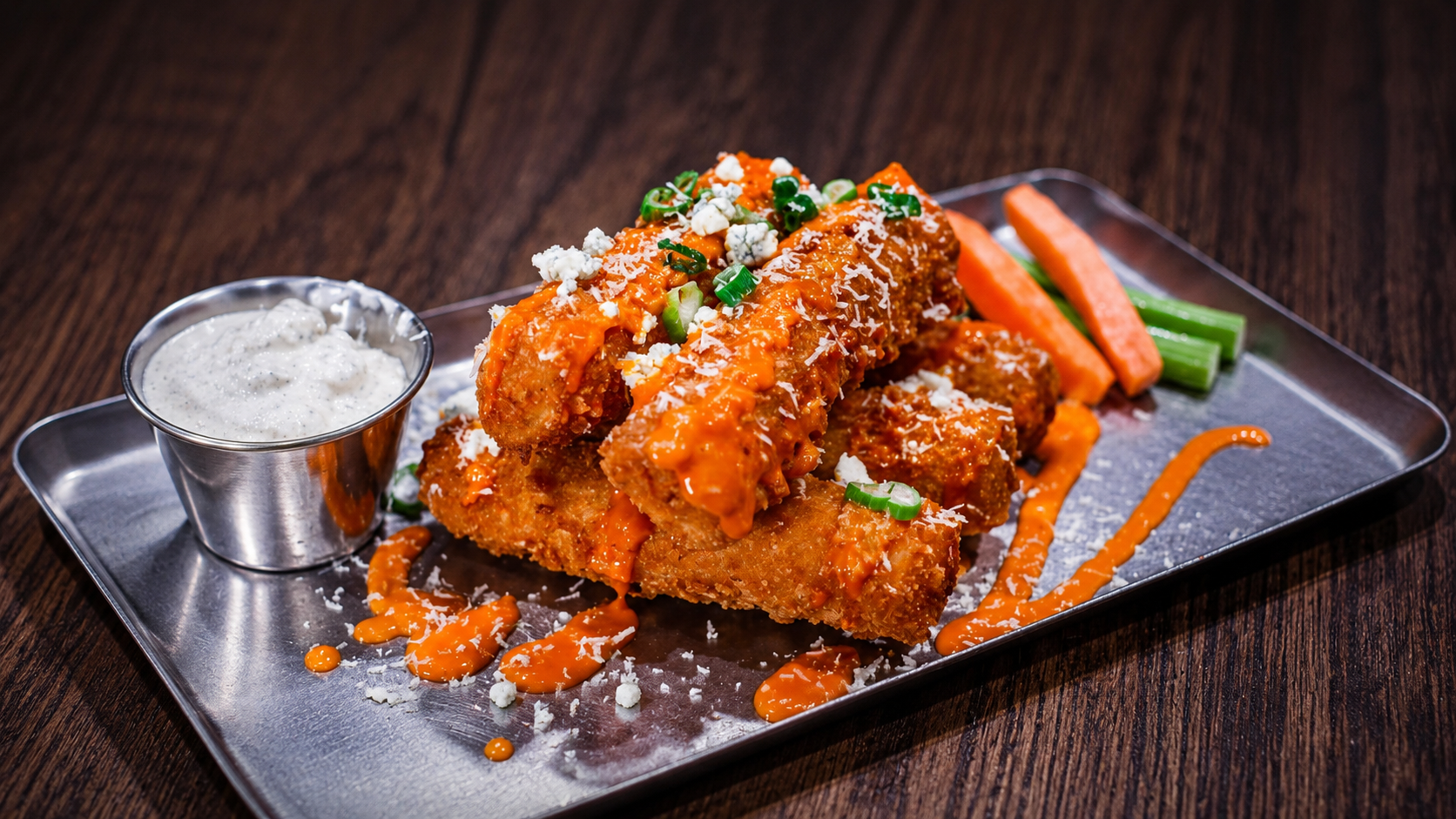 Fried chicken tenders topped with orange sauce, green onions, and shredded cheese on a metal tray, with a side of ranch dressing and celery and carrot sticks in the background.