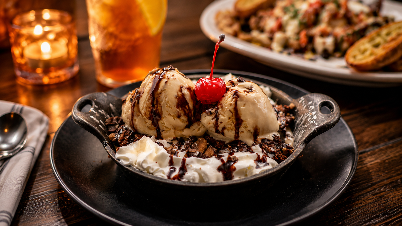 A dessert with three scoops of vanilla ice cream topped with chocolate syrup and a maraschino cherry, served in a dark bowl with whipped cream, chocolate shavings, and cookie crumbles, on a black plate. In the background, a glass of iced tea with lem