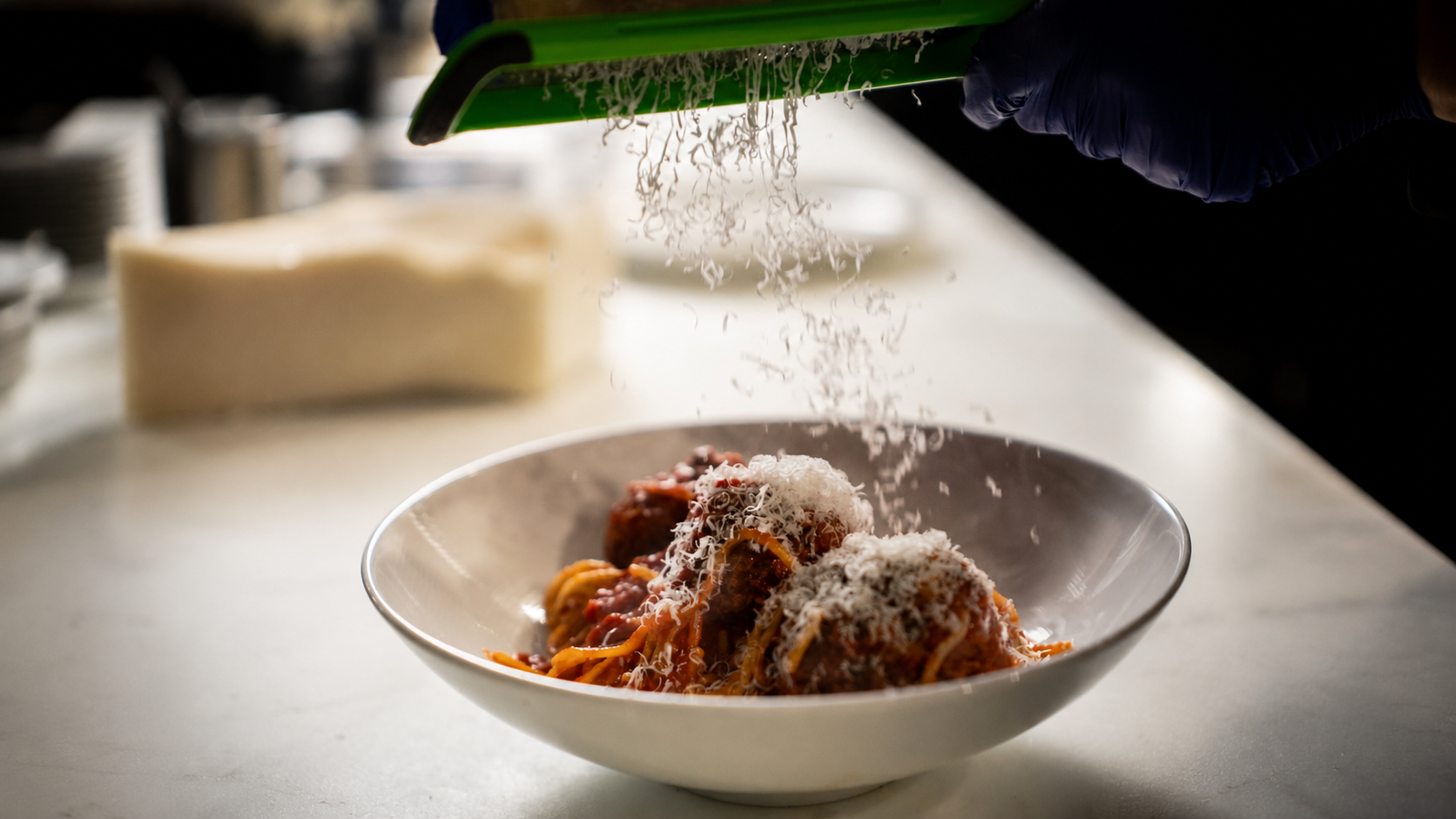 A person wearing gloves sprinkling grated cheese over a bowl of spaghetti and meatballs.