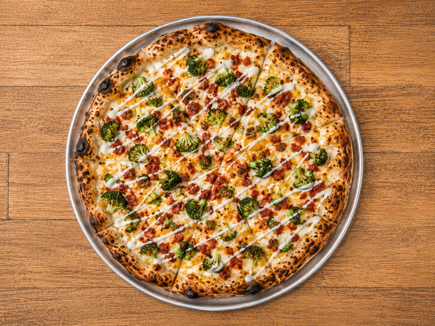 Top-down view of a pizza with broccoli, bacon bits, and white sauce drizzled on top, on a metal tray on a wooden table.