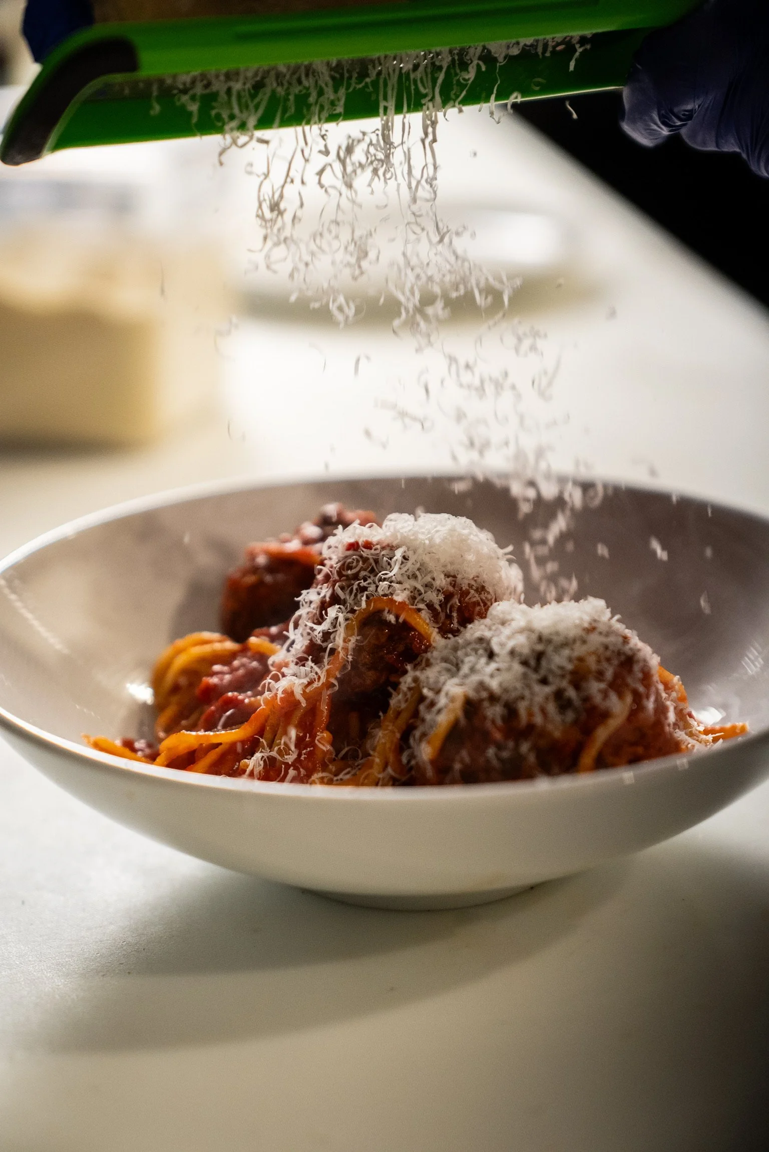 A white bowl of spaghetti with tomato sauce, topped with grated cheese, with some meatballs visible, on a white surface. The image is taken from a low angle, showing a green device or tool in the background, with a person's hand wearing a dark glove in the top right corner.