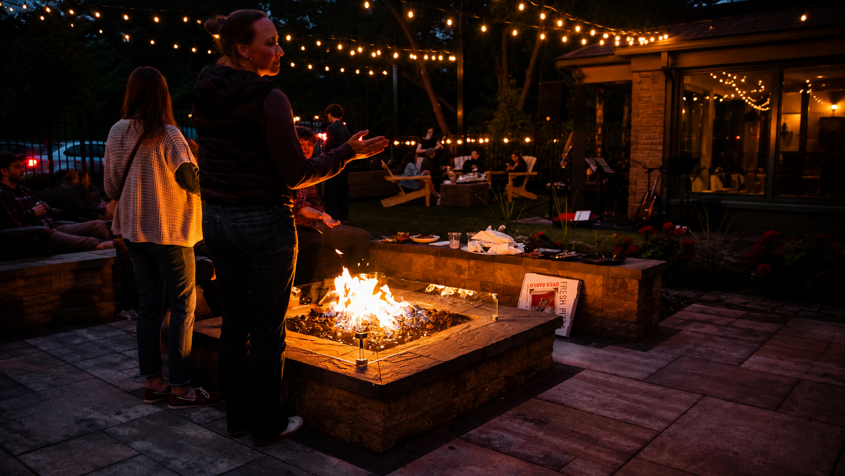 People gathered around a backyard fire pit during an evening outdoor gathering, with string lights overhead and a house in the background.
