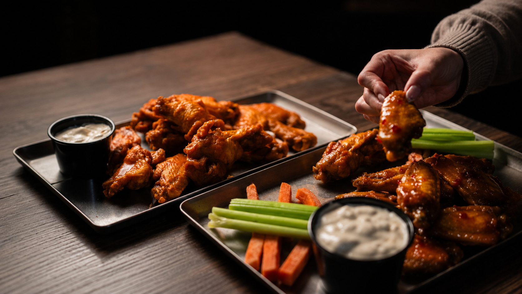Two black trays with buffalo wings, celery sticks, carrot sticks, and dipping sauces on a wooden table. A person is picking up a wing from the tray on the right.