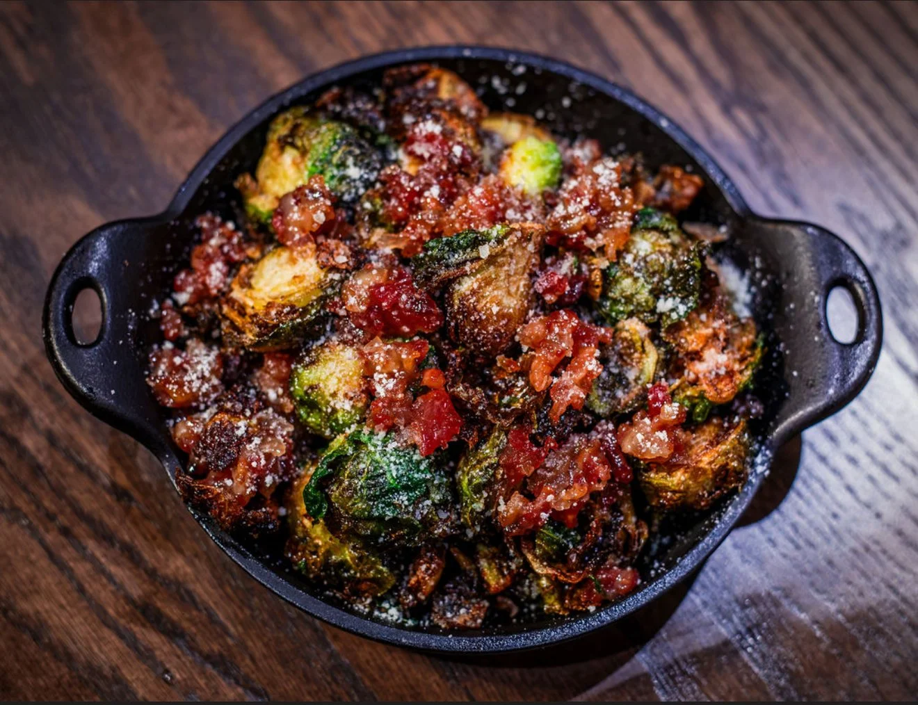 A black cast iron skillet filled with roasted Brussels sprouts topped with tomato sauce and grated cheese, on a wooden table.