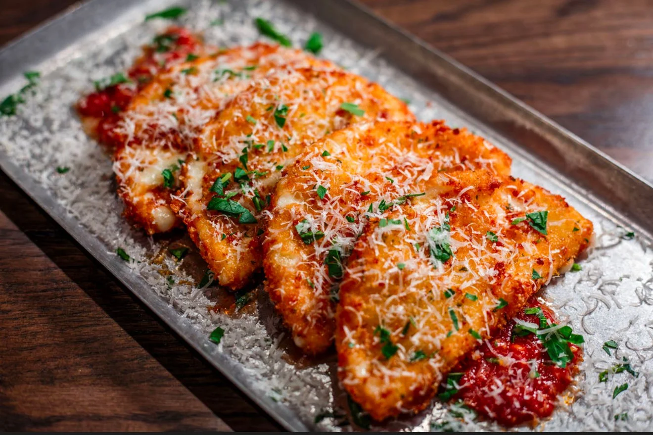 Three crispy breaded chicken cutlets with tomato sauce, shredded cheese, and chopped herbs on a metal tray