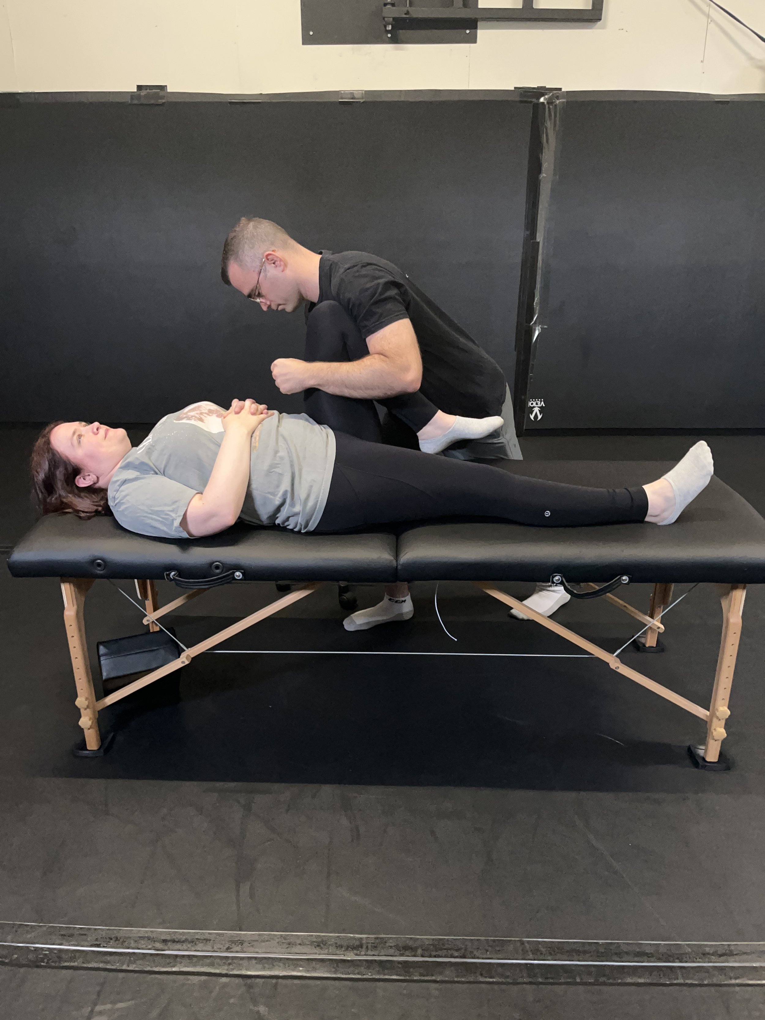 A woman lying on a massage table with her eyes closed and hands clasped on her chest, as a man performs an osteopathic treatment in a room with a dark background and black flooring.