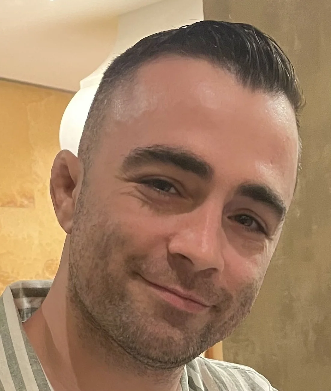 Joe, smiling with short dark hair and a lightly stubbled face, indoors against a yellow wall and a white lamp in the background.