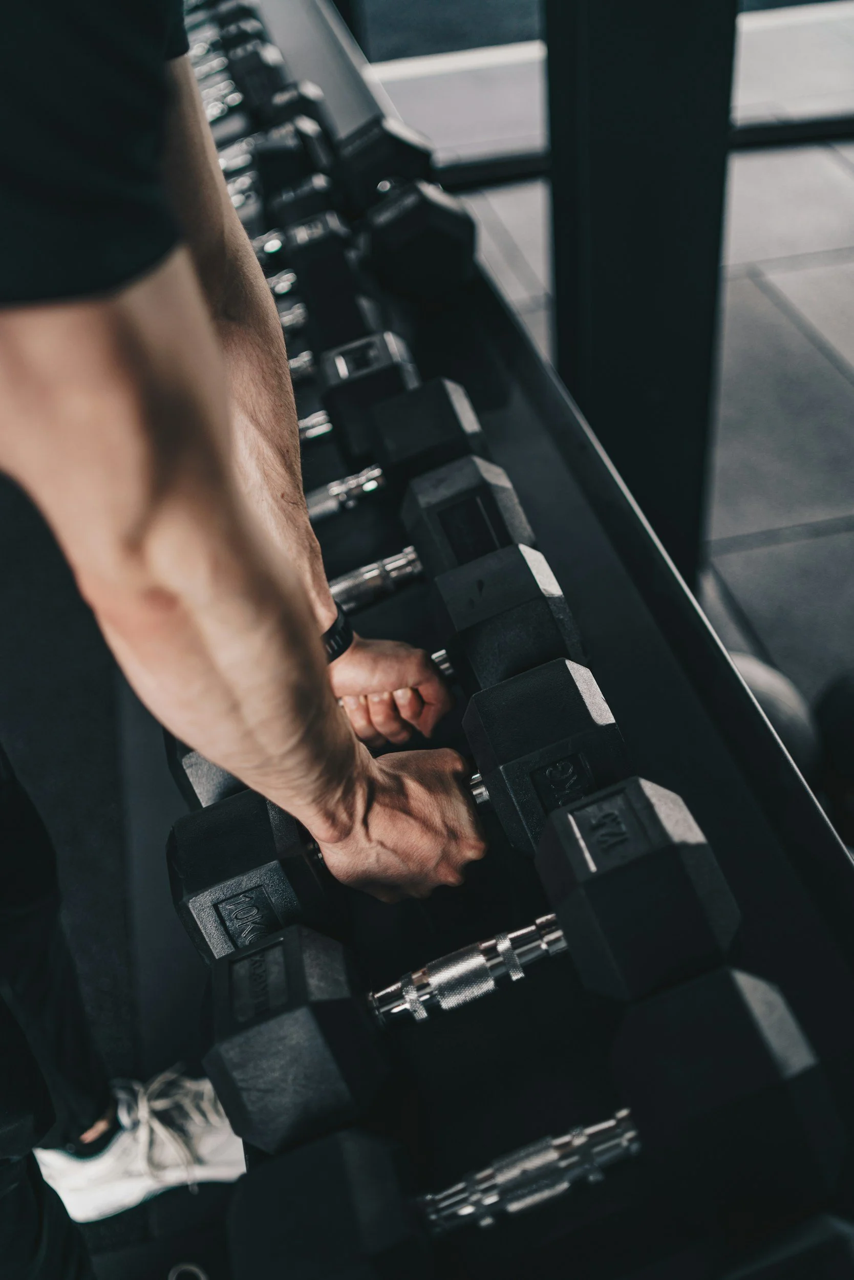 Close-up of a person's arm and hand gripping a dumbbell during a workout, with a row of other black dumbbells on a rack in a gym.