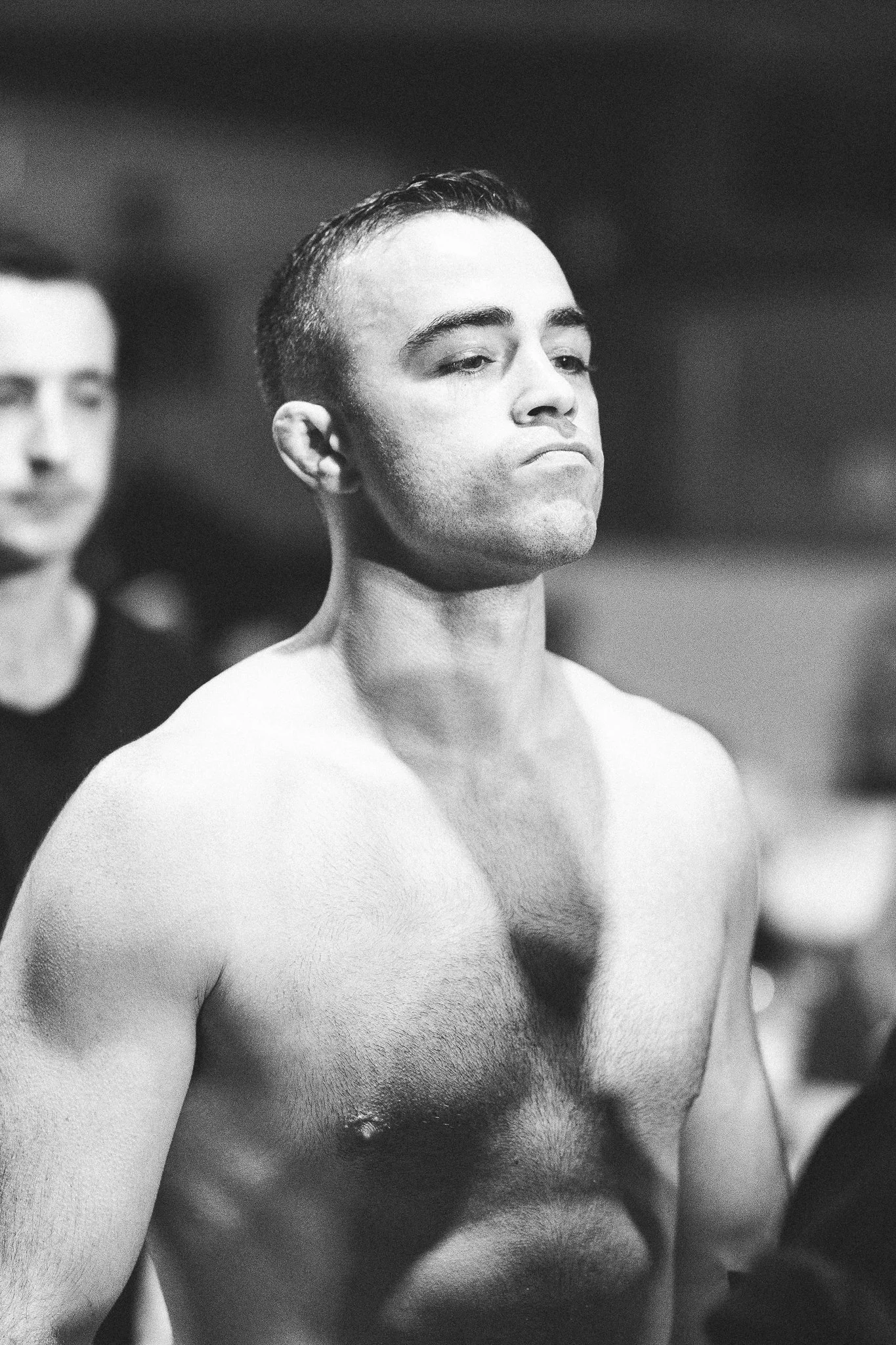 Black and white photograph of Joe, shirtless with short hair and a serious expression, standing with his head slightly turned.
