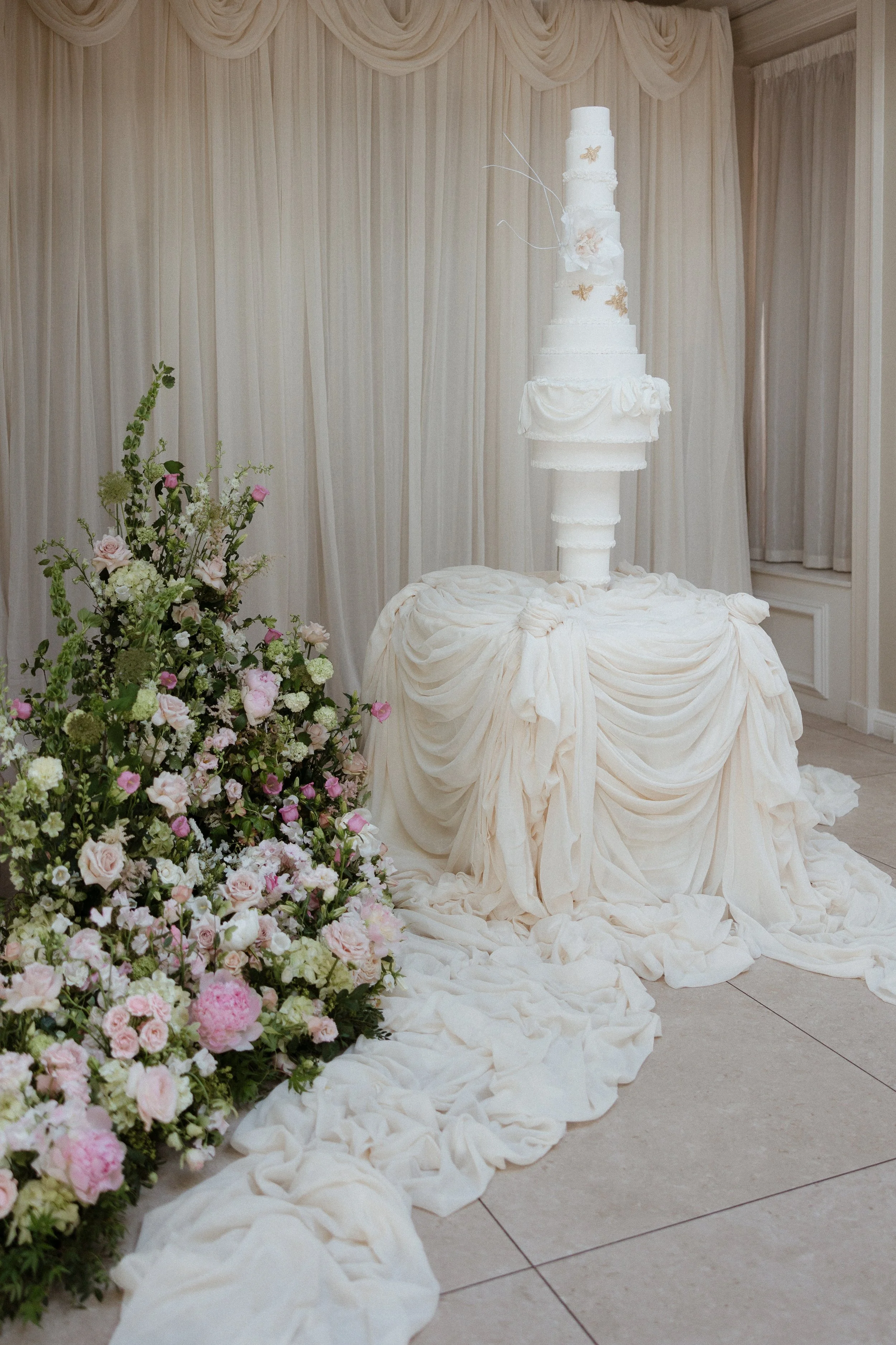 A tall, white wedding cake with gold butterfly decorations and floral accents, displayed on a draped fabric-covered table at a wedding reception.