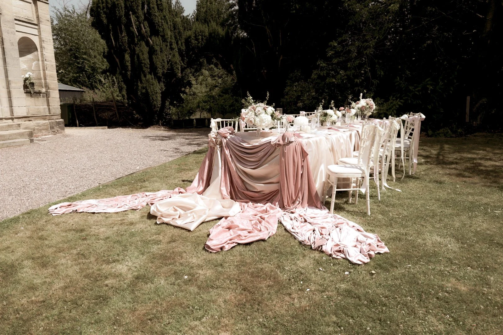 Elegant outdoor wedding table setup with pink and white draped fabrics, floral arrangements, and white chairs on grass, near a stone building and trees.