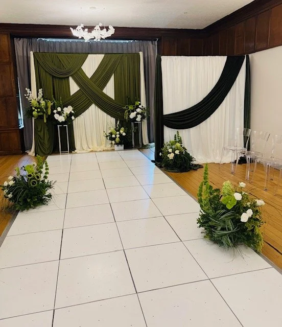 Decorated indoor event space with dark green, black, and white drapes, floral arrangements with greenery and white flowers, and a white tiled floor.