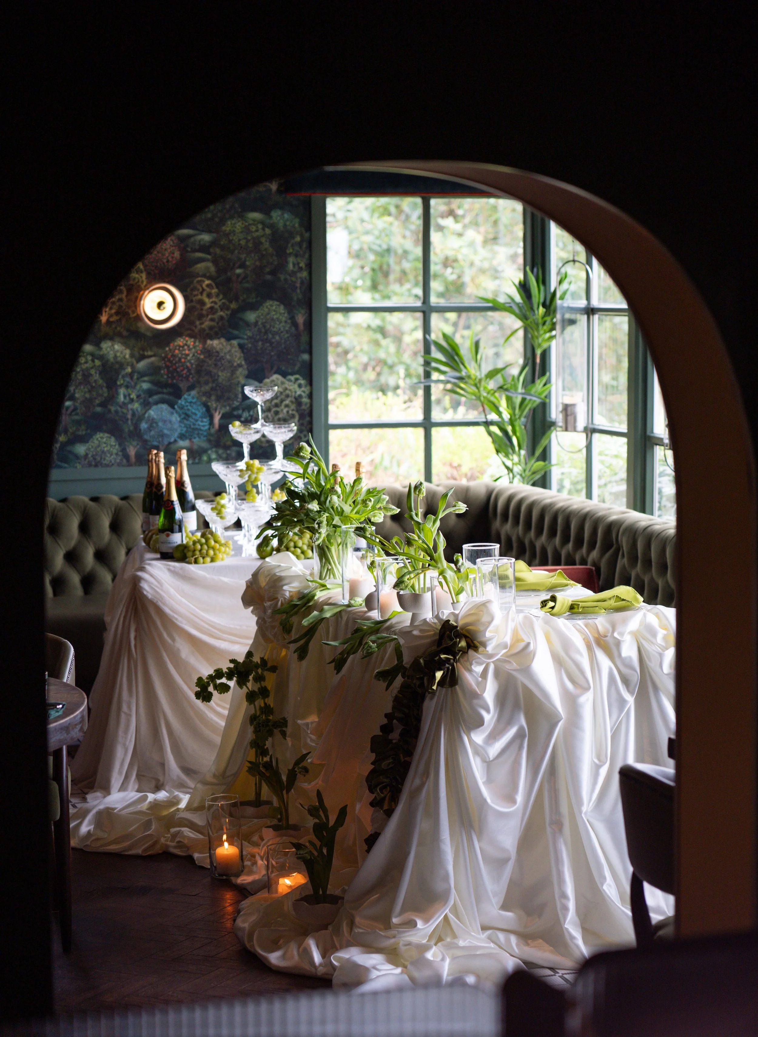 A decorated dining table with a white tablecloth, greenery, bottles of champagne, wine glasses, a multi-tiered glass stand, candles, and lush plants near a large window, viewed through an arched opening.
