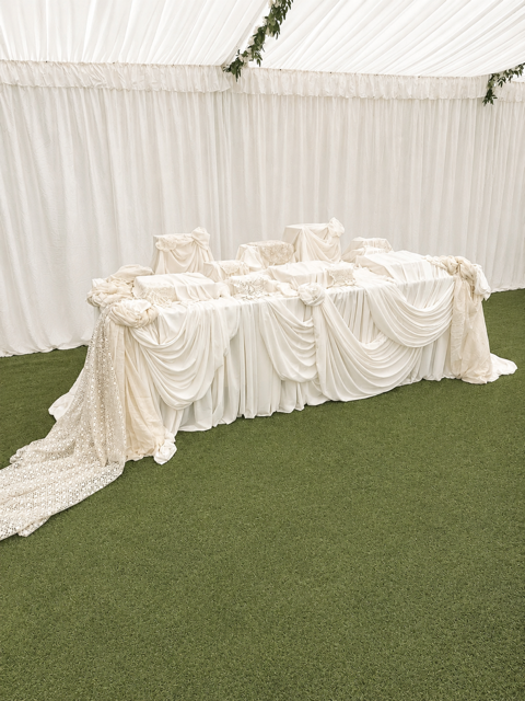 Wedding head table draped in white fabric with chairs and a backdrop of white curtains, decorated for a celebration.