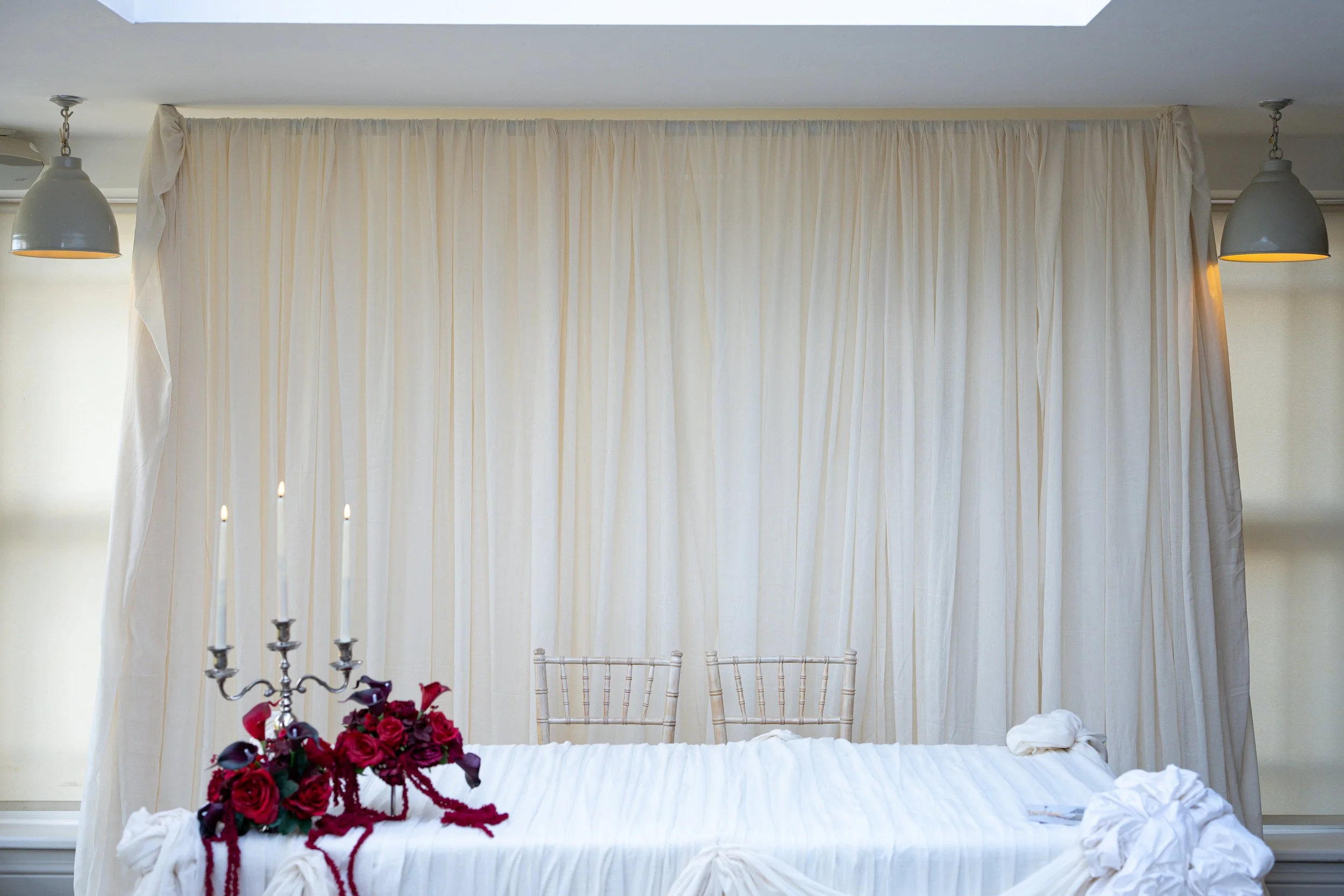 A bedroom with a white bed, beige curtains, two hanging gray lamps, a white chair, a candelabra with candles, and a bouquet of red and purple flowers.
