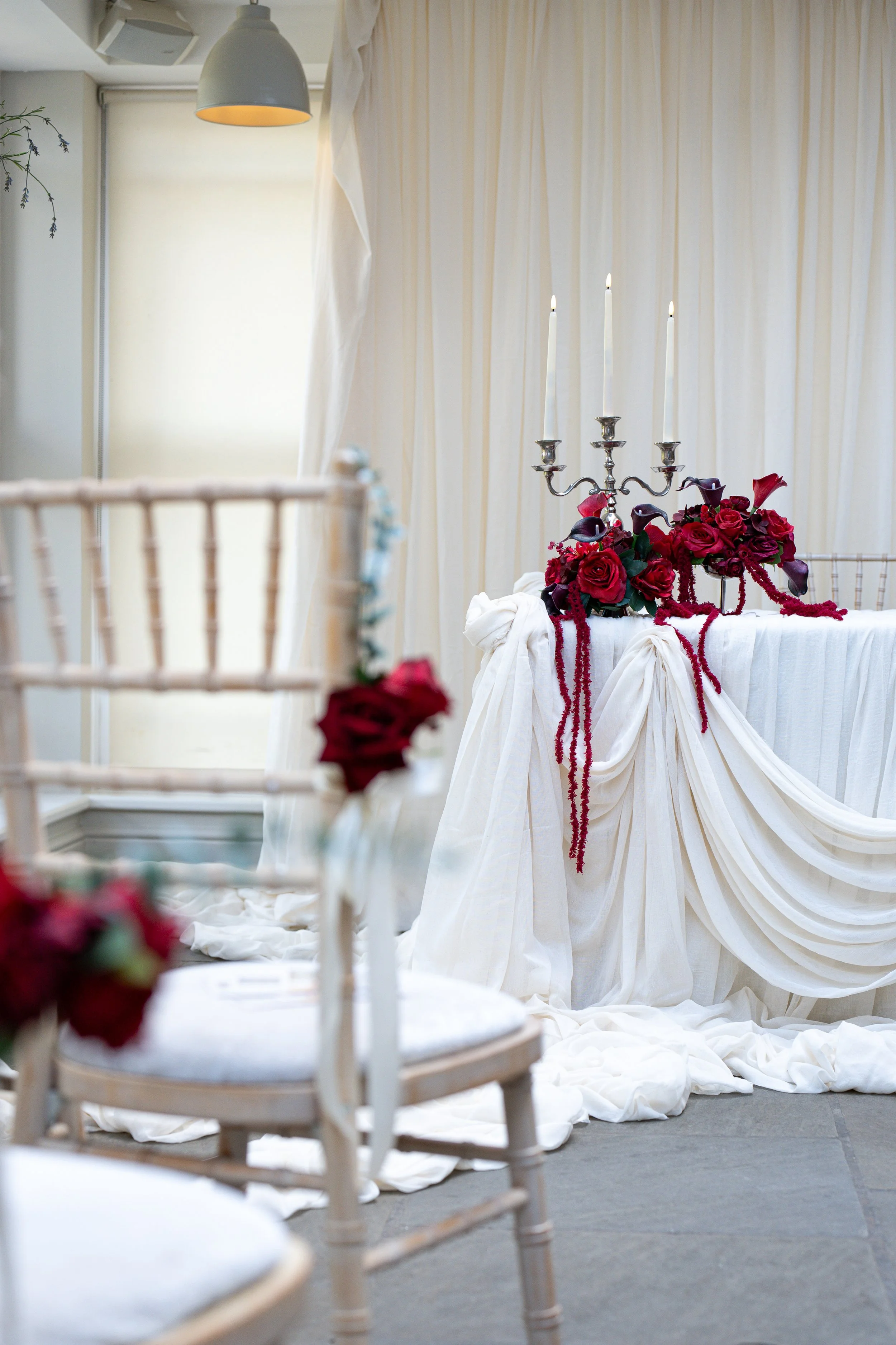 A wedding or formal event setup with a white draped table decorated with a silver candelabrum holding four lit candles and a bouquet of red and purple roses. The background features a cream-colored curtain and a white ceiling with a hanging pendant light.