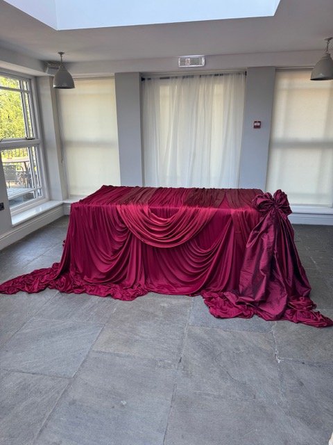 A table covered with a red satin cloth with draped and ruched fabric, tied with a bow on the right side, located in a room with large windows and white curtains.